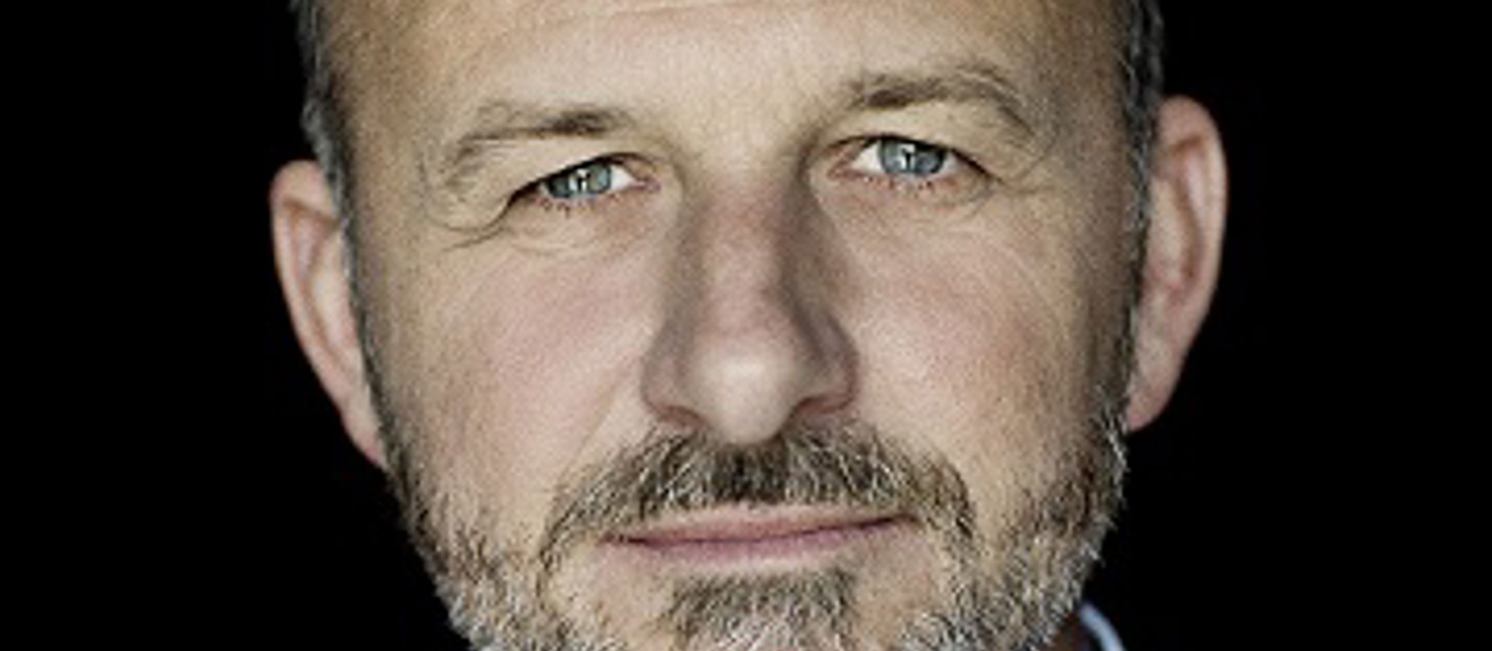 Instruktøren Nikolaj Cederholm iført hvid åbenstående skjorte med blå milde øjne og lukkede læber smiler svagt.