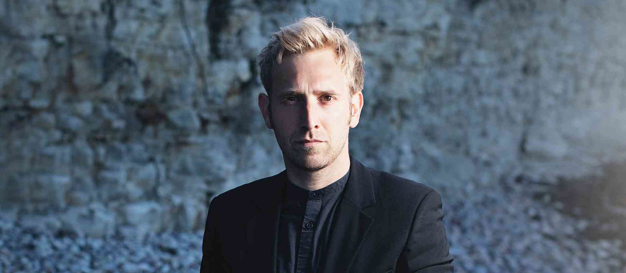 Akkordeon-virtuosen Bjarke Mogensen med lyst hår og brune intense øjne iført sort skjorte og jakke foran sløret blå baggrund.