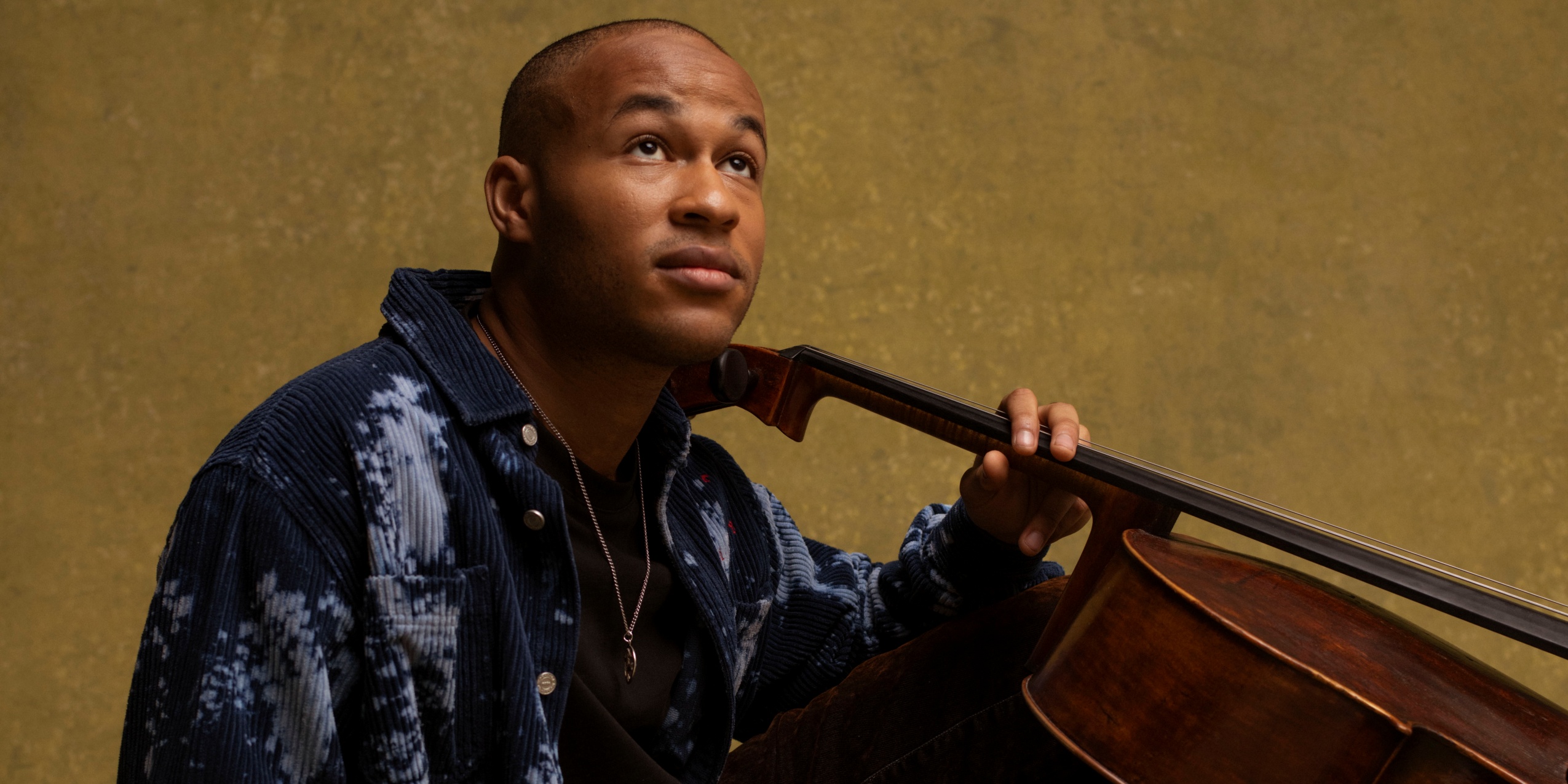 Mand siddende på gulv med en cello foran mørkegul væg 