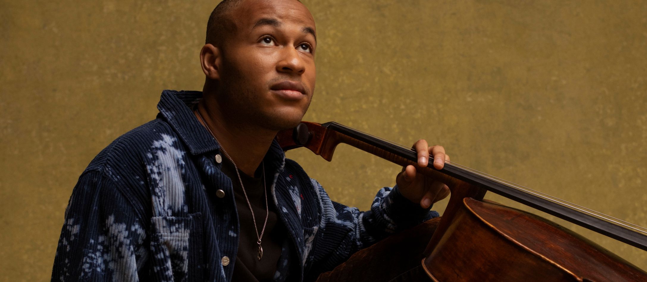 Mand siddende på gulv med en cello foran mørkegul væg